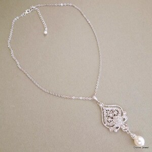 A silver-colored necklace with an ornate, filigree-style pendant encrusted with clear crystals and a single pearl drop, displayed on a gray background.
