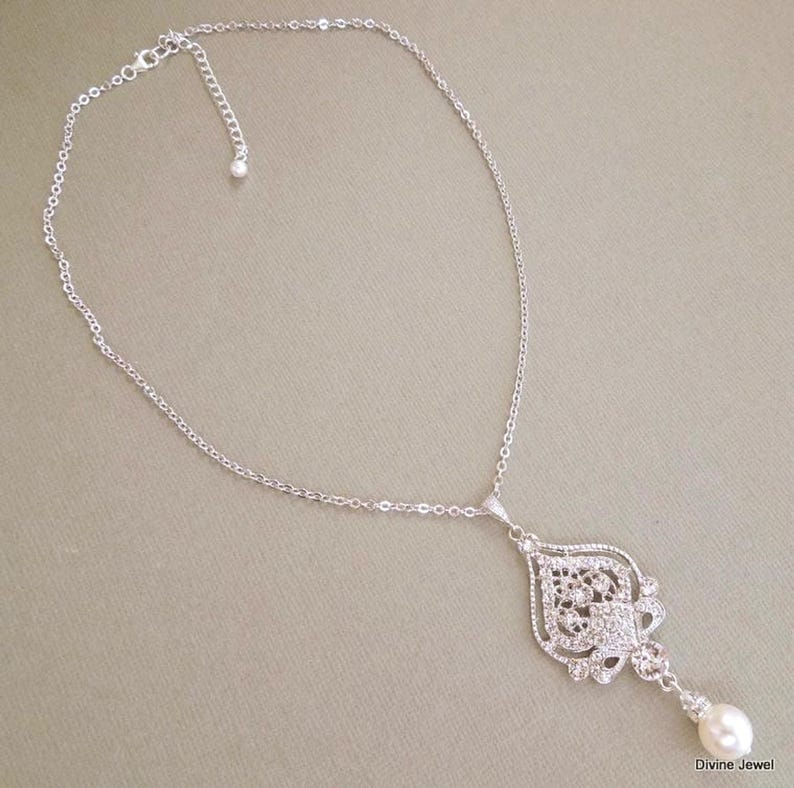 A silver-colored necklace with an ornate, filigree-style pendant encrusted with clear crystals and a single pearl drop, displayed on a gray background.