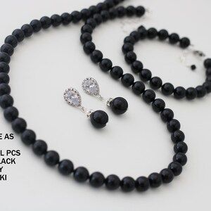 K&ouml;nnte beinhalten: Ein Schmuckset bestehend aus einer Halskette und einem Armband aus runden schwarzen Perlen, begleitet von einem Paar Ohrh&auml;nger mit klaren tropfenf&ouml;rmigen Steinen und schwarzen Perlenakzenten. Text: PURCHASE AS SET OR INDIVIDUAL PCS MYSTIC BLACK PEARLS BY SWAROVSKI.