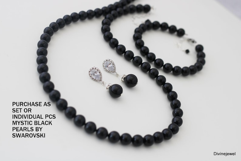 K&ouml;nnte beinhalten: Ein Schmuckset bestehend aus einer Halskette und einem Armband aus runden schwarzen Perlen, begleitet von einem Paar Ohrh&auml;nger mit klaren tropfenf&ouml;rmigen Steinen und schwarzen Perlenakzenten. Text: PURCHASE AS SET OR INDIVIDUAL PCS MYSTIC BLACK PEARLS BY SWAROVSKI.