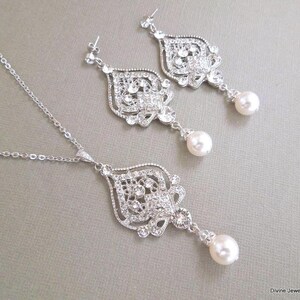 A silver-colored necklace and matching earrings, each with an ornate, filigree-style pendant encrusted with clear crystals and a single pearl drop, displayed on a gray background.