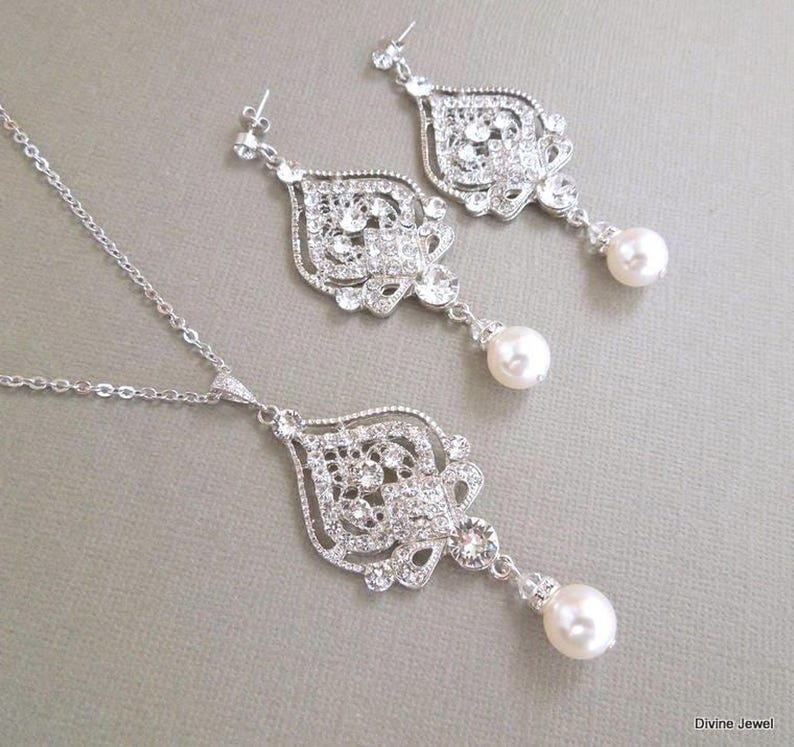 A silver-colored necklace and matching earrings, each with an ornate, filigree-style pendant encrusted with clear crystals and a single pearl drop, displayed on a gray background.