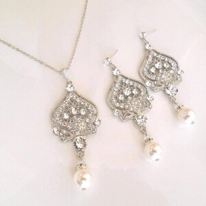 A silver-colored necklace and matching earrings, each with an ornate, filigree-style pendant encrusted with clear crystals and a single pearl drop, displayed on a white background.
