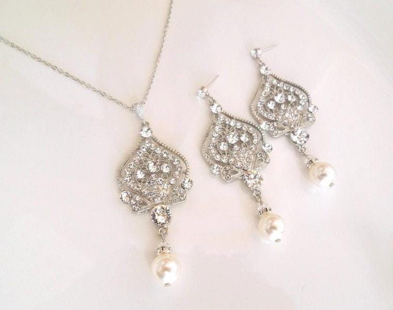A silver-colored necklace and matching earrings, each with an ornate, filigree-style pendant encrusted with clear crystals and a single pearl drop, displayed on a white background.