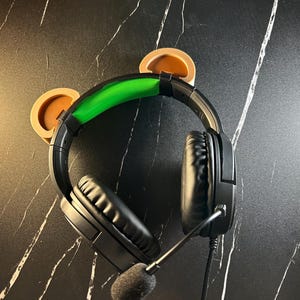 May include: Black gaming headset with green accents and brown earcup covers. The headset has a microphone attached.