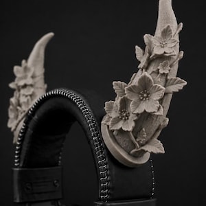 May include: A pair of matte gray, horn-shaped attachments mounted onto a black headset. Each horn features a detailed, sculpted relief of five-petaled flowers and leaves winding around the surface, creating a textured, floral aesthetic.
