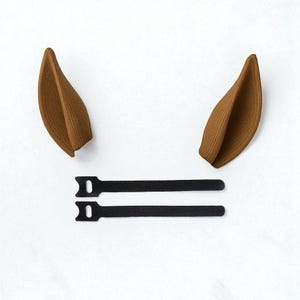 May include: Two brown, textured, ear-shaped accessories and two black, adjustable straps, against a white background. The ear shapes are curved and have a ridged texture. The straps are flat and rectangular with a loop at one end.