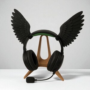 May include: Black headphones with a microphone, featuring black wings attached to the headband. The headphones rest on a wooden stand. The headband has a green accent.
