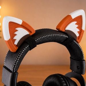 May include: A pair of black over-ear headphones featuring detachable 3D-printed fox ears attached to the headband. The fox ears are colored in orange, white, and dark brown with a textured finish. The headband has visible white stitching along the edges.