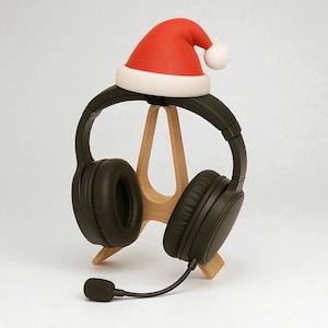 Può includere: Cuffie nere over-ear con microfono, appoggiate su un supporto in legno. Un piccolo cappello da Babbo Natale rosso con bordo bianco e pompon è posto sopra, suggerendo un tema festivo. Le cuffie sono posizionate su uno sfondo bianco.