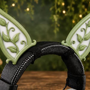 May include: A black headband featuring two pointed, upright ears with a light green frame and a white center. Each ear is decorated with a raised, sculpted green vine pattern with leaves and swirling stems attached to the front of the structure.