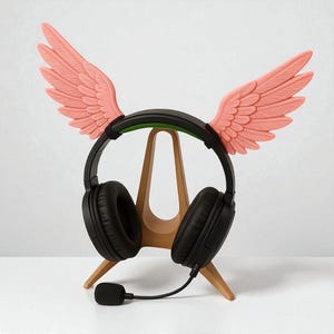 Puede incluir: Auriculares negros con micrófono, apoyados en un soporte de madera. Los auriculares están adornados con alas rosas en forma de pluma. El soporte es de color marrón claro y el fondo es blanco.