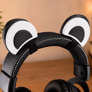 May include: Black over-ear headphones featuring a pair of detachable, semi-circular animal ears attached to the top headband. The ears are white with black inner accents, and the headband is finished with a textured black material and silver stitching.