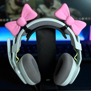 May include: Light grey gaming headphones with pink bow decorations. The headphones have grey earcups and are displayed on a black stand. A computer monitor and keyboard are visible in the background.