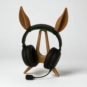 May include: A wooden headphone stand with bunny ear attachments. The stand supports black headphones with a microphone. The stand is made of light brown wood and has a simple, minimalist design.