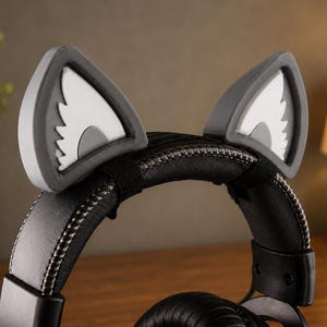 May include: A pair of gray and white cat ear attachments secured to the top of black headphones. The ears feature a triangular gray frame with white inner sections and a jagged, stylized design. The headphones have a padded black headband with visible stitching.