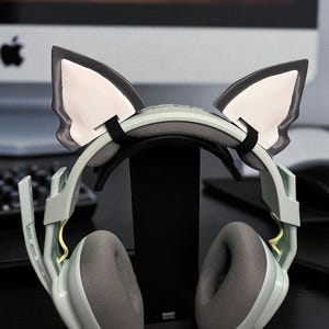 May include: A pair of gray and black gaming headphones with cat ear attachments. The headphones have gray earcups and a black headband. The cat ears are black with white inner ears. The headphones are resting on a black stand.