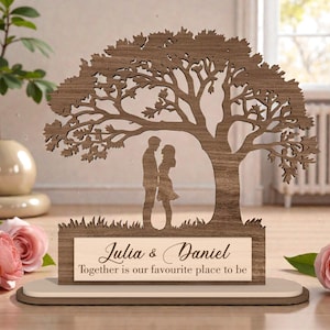 Può includere: Silhouette in legno di un albero con una coppia sotto. La base presenta i nomi "Julia & Daniel" e la frase "Together is our favourite place to be". L'albero e la base sono marrone chiaro.
