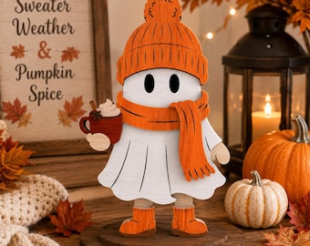 May include: A white wooden ghost figurine wearing an orange knit hat and matching scarf, holding a small red mug with white whipped cream. The ghost stands on a circular wooden base and is wearing orange boots, featuring a simple design.
