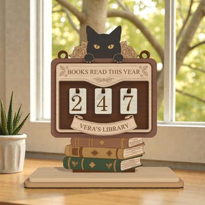 Può includere: Un contatore di libri in legno con un gatto nero in cima, che mostra "247" libri letti. Il contatore recita "BOOKS READ THIS YEAR" e "Vera's Library". Il contatore è posto sopra una pila di libri, con una piccola pianta in vaso sullo sfondo.