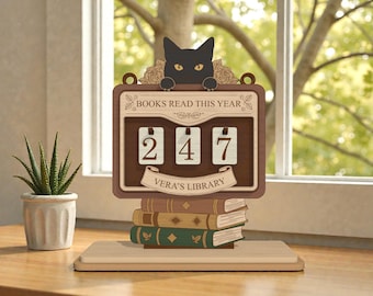 May include: A wooden book counter with a black cat on top, displaying "247" books read. The counter reads "BOOKS READ THIS YEAR" and "Vera's Library". The counter sits atop a stack of books, with a small potted plant in the background.