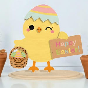 Puede incluir: Decoración de Pascua de madera con un pollito amarillo saliendo de una cáscara de huevo agrietada. El pollito sostiene un cartel que dice "Happy Easter!" y una cesta con huevos decorados. Dos cactus en macetas flanquean la decoración.