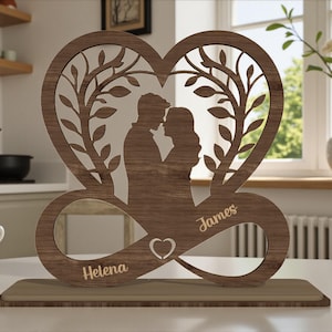 May include: A wooden heart-shaped decorative piece featuring a couple embracing within a tree design. The names "Helena" and "James" are engraved on the infinity symbol base. The piece is set on a rectangular wooden stand.