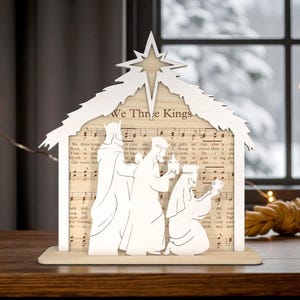 May include: A wooden Christmas ornament showing the Three Kings, crafted from light wood. The design includes a star, a roof, and the figures of the kings, set against a backdrop of sheet music. The text "We Three Kings" is visible.