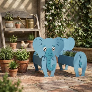 May include: Two light blue elephant-shaped planters with a rectangular trough for plants. The planters have large ears, a trunk, and a tail. The planters are on a brick patio, with other potted plants in the background.