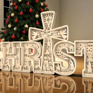 May include: A wooden Christmas decoration spelling out the word "CHRIST" with a cross. The letters and cross are intricately carved with religious scenes. A Christmas tree with red and gold ornaments is in the background.