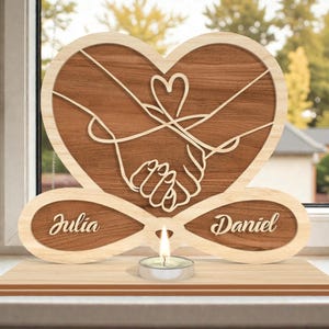 May include: A wooden heart-shaped decoration with a carved design of two hands holding each other. The names "Julia" and "Daniel" are engraved on the base. A small candle sits in front of the heart. The wood has a light and dark brown color.