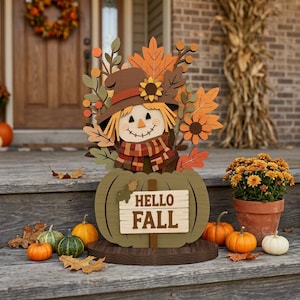 May include: A wooden scarecrow decoration featuring a brown hat, a plaid scarf, and autumn leaves, sitting atop a green pumpkin base with a sign that reads HELLO FALL. The cutout is displayed on a dark circular wooden stand on a porch.
