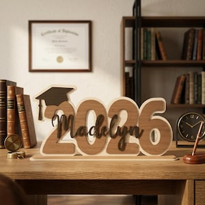 May include: Wooden graduation decoration featuring the year 2026 and the name Madelyn, with a graduation cap. The numbers and name are in a light brown color, with a darker brown graduation cap. The decoration is on a wooden base.