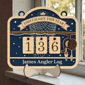 Puede incluir: Registro de pesca de madera con el texto "FISH CAUGHT THIS YEAR" y "James Angler Log". El letrero tiene un fondo azul marino con un diseño de red de pesca y carrete. Se muestran los números "136".