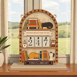 Può includere: Decorazione in legno a forma di libreria con impronte di zampe, libri e un gatto che dorme. Il testo "BOOKS READ THIS YEAR" è visualizzato sopra il numero "247". Sono visibili anche una tazza di caffè, una caffettiera a versare e il testo "VERA'S LIBRARY".