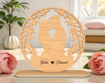 Può includere: Un ornamento in legno raffigurante la silhouette di una coppia che si bacia, incorniciata in un cerchio con intagli floreali. I nomi "Julia" e "Daniel" sono incisi sotto, separati da un cuore. Base rettangolare.