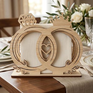May include: Wooden wedding ring-themed centerpiece. The design features two interlocking rings, one with a diamond-shaped accent and the other with a crown. A heart shape is cut out in the center. The base has a leaf design.