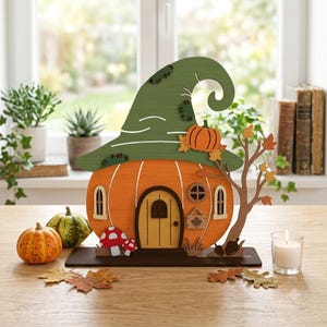 May include: A wooden pumpkin-shaped house decoration featuring a large green witch hat roof, a central arched door, two small windows, and a circular window. The design includes a brown tree with orange leaves, red and white mushrooms, and a small sign.