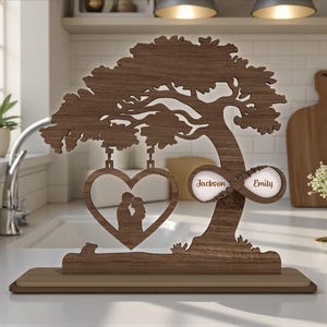 May include: A wooden tree ornament with a heart-shaped cutout showing a couple's silhouette. The tree features an infinity symbol with the names "Jackson" and "Emily". The base is light brown.