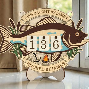Puede incluir: Letrero de madera con forma de pez con el texto "FISH CAUGHT BY JAMES" y "HOOKED BY JAMES". El letrero muestra el número "136" en un contador. Presenta un diseño de pez con colores marrón, azul y beige, junto con un ancla y una fogata.