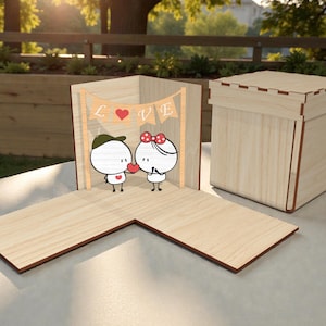 May include: A wooden box with a hinged lid and an open display area. The display features a cartoon couple holding a heart, with a banner that says "LOVE". The box is made of light-coloured wood with darker edges.