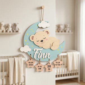 Può includere: Cartello in legno con nome del bambino raffigurante un orsetto addormentato su una luna blu a mezzaluna, stelle gialle e nuvole bianche. Il nome "Finn" è scritto in corsivo. Include etichette a forma di stella con i dettagli della nascita.
