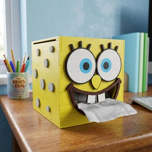 Puede incluir: Caja de pañuelos de madera amarilla con la cara de un personaje de dibujos animados con ojos grandes, una gran sonrisa y una lengua. La caja tiene forma cuadrada con detalles circulares y un pañuelo sale de la boca. El diseño está inspirado en un personaje animado popular.