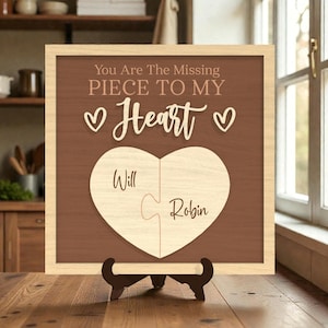 May include: Wooden framed sign with the text "You Are The Missing Piece To My Heart" and two heart-shaped puzzle pieces. The names "Will" and "Robin" are inscribed on the puzzle pieces. The sign is displayed on a wooden stand.