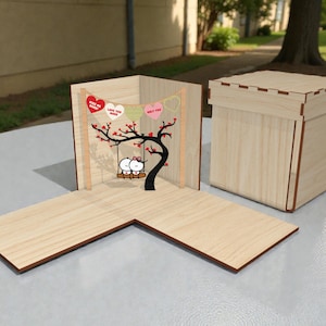 May include: A wooden pop-up card with a tree and swing design, featuring two cartoon figures. The card includes heart-shaped banners with text. A separate wooden box is also present, with a matching design. The card is open, revealing the interior.