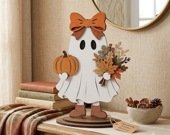 May include: A white wooden ghost cutout decoration featuring a large orange bow on its head, wearing brown boots, and holding an orange pumpkin in one hand and a bouquet of autumn-colored leaves and berries in the other, standing on a round base.