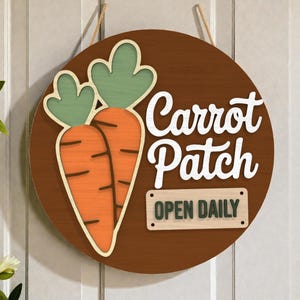 Puede incluir: Un letrero redondo de madera marrón con dos zanahorias naranjas y hojas verdes. Las palabras "Carrot Patch" están en escritura blanca, y un letrero más pequeño debajo dice "OPEN DAILY".
