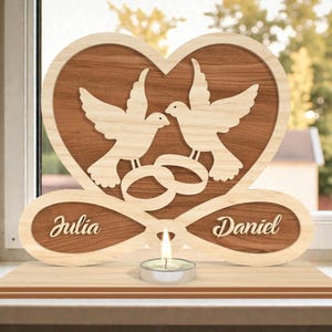 Op de afbeelding: Houten bruiloftsdecoratie in hartvorm met twee duiven, in elkaar grijpende ringen en de namen "Julia" en "Daniel". Een kleine kaars staat eronder. Het ontwerp is gemaakt van licht en donker hout.