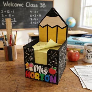 Può includere: Un organizer da scrivania a forma di matita in legno nero con interno giallo e design a lavagna. L'organizer ha il nome "Mrs. Horton" in lettere colorate. All'interno c'è una pila di post-it gialli.