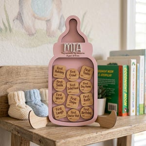 May include: A pink wooden baby bottle-shaped keepsake with the name "MIA" and the text "Tiny moments, big memories, A year of firsts." Inside are wooden tokens for recording baby's milestones. Beside it are baby booties and books.